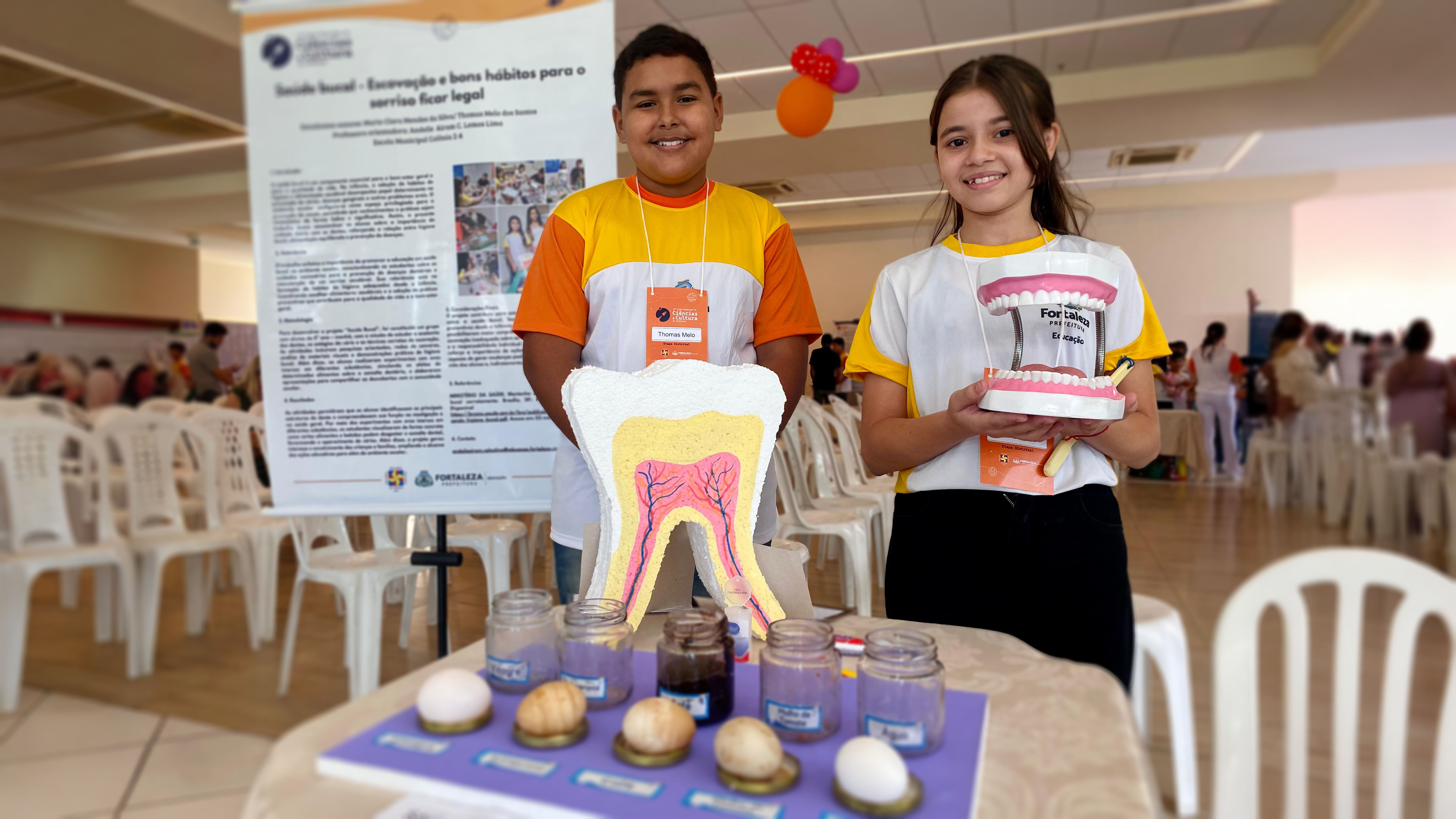 A imagem mostra o projeto desenvolvido pelos alunos Maria Clara Mendes e Thomas Melo que utiliza ovos imersos em líquidos como refrigerante e vinagre para simular os efeitos da má escovação dental